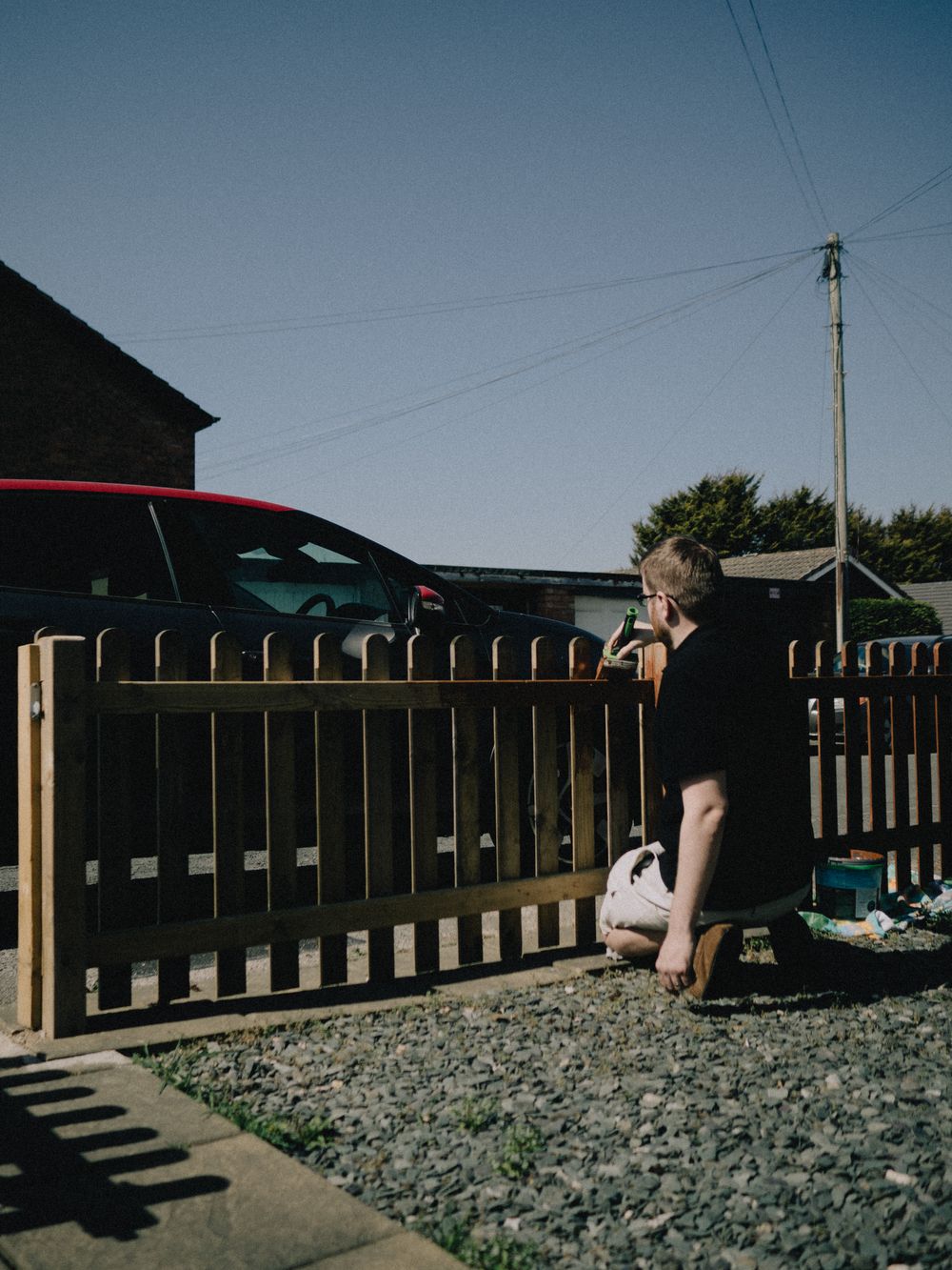 A person crouched down painting a timber picket fence in bright sun